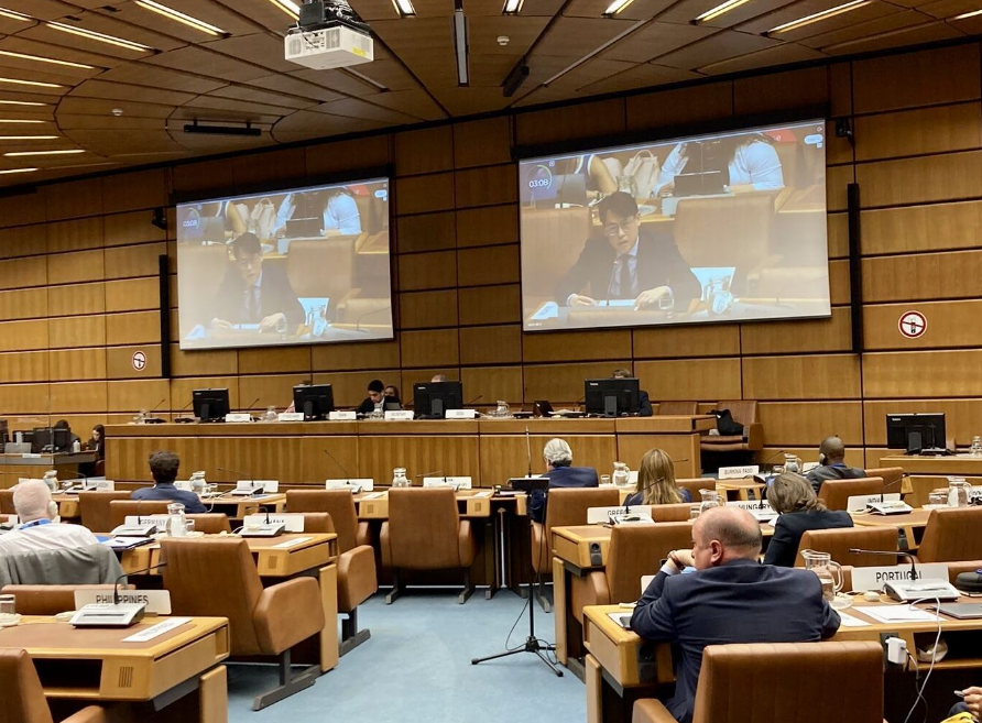 Conference room with attendees seated at desks, facing a large screen displaying the speaker and panels with computers.